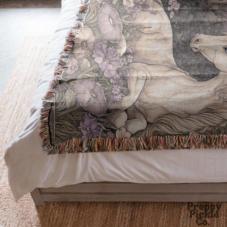 Op de afbeelding: Een geweven sprei met een gedetailleerde illustratie van een wit paard met paarse bloemen en bladeren. De deken heeft een gefranjerde rand met een mix van kleuren. Het paard ligt en het ontwerp heeft een vintage esthetiek.