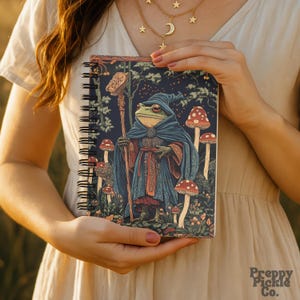 Peut inclure: Cahier à spirales avec une illustration fantastique d'un magicien grenouille en cape bleue, tenant un bâton et entouré de champignons. La couverture présente une scène de forêt bleu foncé. Le cahier est tenu par une personne.