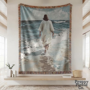 May include: Woven tapestry showing a figure in a white robe walking into the sea. The artwork features a calm seascape with shades of blue and grey, and a fringed border. The tapestry is a decorative wall hanging.