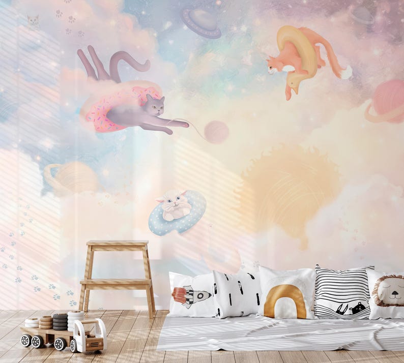 May include: A whimsical nursery wall mural featuring a pastel pink, blue, and yellow color scheme. The mural depicts three cats flying through the air with planets and clouds in the background. The cats are wearing a donut, a ring, and a cloud.