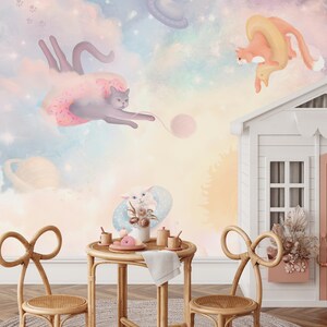 May include: A whimsical children's room with a pastel pink, blue, and yellow wall mural featuring cats and clouds. The mural is accented with a white playhouse with a pink door and a sign that says "Love". Two wicker chairs with bow-shaped backs are placed in front of a small wooden table with a white tablecloth and a plate of cookies.