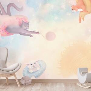 May include: A nursery wall mural with a pastel pink, blue, and yellow color scheme. The mural features three cats playing with a ball of yarn. One cat is wearing a pink tutu and flying through the air. The mural is perfect for a child's room.