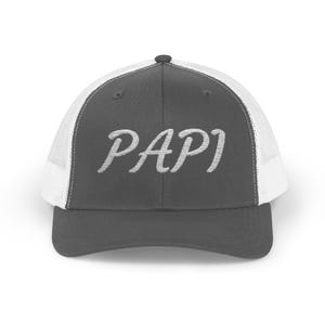 May include: A grey trucker cap with a white mesh back and a curved peak. The word "PAPI" is embroidered in white script across the front. The cap features a black button on the crown.
