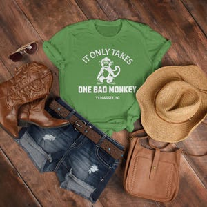 May include: Green t-shirt with a white monkey graphic and the text "It only takes one bad monkey Yemassee, SC". The shirt is paired with denim shorts, brown boots, a straw hat, and a brown leather crossbody bag.