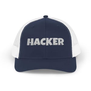 Puede incluir: Gorra de camionero azul marino con parte trasera de malla blanca y visera curva. La palabra "HACKER" está bordada en una fuente gris claro texturizada en la parte delantera. Estilo clásico y casual.