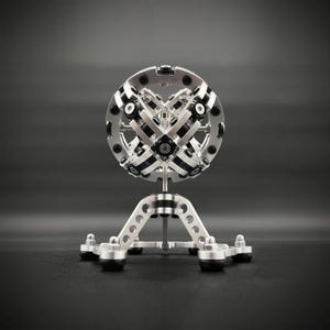 Pode incluir: Uma escultura de metal prateado com um design geométrico complexo. A escultura é composta por múltiplas peças que se entrelaçam e giram livremente. Está montada em uma base de tripé.