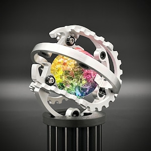 Può includere: Una scultura meccanica color argento con una sfera cristallina multicolore al centro. La sfera è racchiusa in una complessa disposizione di ingranaggi ed elementi metallici curvi, montata su una base cilindrica nera.