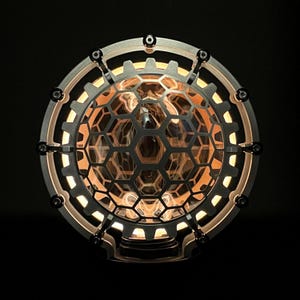 May include: A round, metal lamp with a honeycomb-shaped, translucent glass shade. The lamp has a gear-like design around the edge and is lit from within, casting a warm glow.