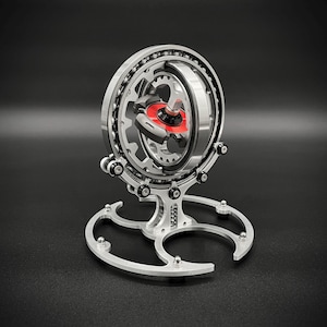 Könnte beinhalten: Eine kinetische Skulptur aus silbernem Metall mit einem roten Mittelstück. Die Skulptur ist auf einem dreizackigen Sockel montiert.