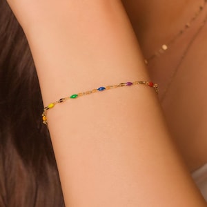 May include: A delicate gold bracelet featuring colorful enamel beads in a rainbow of hues. The bracelet is worn on the wrist and is paired with a gold necklace. The jewelry is a vibrant accessory.