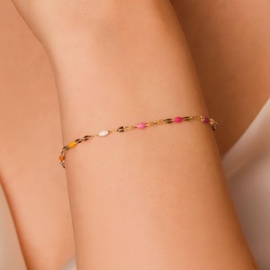 May include: A delicate gold bracelet with colorful, oval-shaped beads. The bracelet features a series of small, vibrant beads in shades of pink, yellow, orange, and white. The jewelry is worn on the wrist.