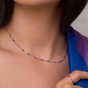 May include: A delicate silver necklace with small, colorful beads in shades of pink and blue. The necklace is worn around the neck and is a simple yet elegant piece of jewelry.