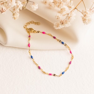 May include: A gold bracelet with a delicate chain design. The bracelet features alternating pink, blue, and purple oval-shaped beads. The bracelet is displayed on a white surface with white flowers in the background.