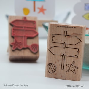 Könnte beinhalten: Holzstempel mit Strandmotiven. Ein Stempel zeigt ein Richtungsschild mit Pfeilen, einer Muschel und einem Seestern. Der andere Stempel hat einen roten Tintenabdruck des gleichen Designs. Die Website www.holzundpoesie.de ist auf dem Stempel aufgedruckt.