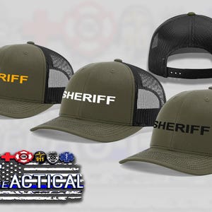 May include: Olive green Sheriff trucker hats with black mesh backs. The word "SHERIFF" is displayed in yellow, white, and black lettering on the front of the hats. The hats have a curved brim and adjustable snapback closure.