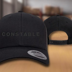 May include: Black mesh back baseball cap with a black brim and a black crown. The cap has the word "CONSTABLE" embroidered in white on the front.