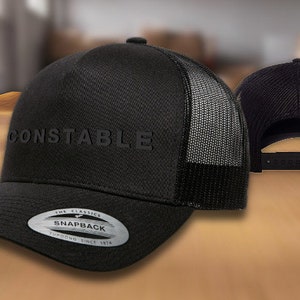 May include: Black mesh trucker hat with a black front panel and the word "CONSTABLE" embroidered in black. The hat has a black snap closure and a silver sticker that says "The Classics Snapback Founded Since 1978".