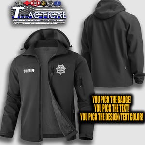 May include: Black softshell jacket with a hood, zipper closure, and multiple pockets. The jacket features the word "SHERIFF" and a badge on the front. The text "YOU PICK THE BADGE! YOU PICK THE TEXT! YOU PICK THE DESIGN/TEXT COLOR!" is also visible.