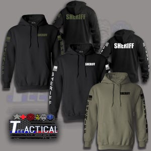 May include: A collection of hooded sweatshirts in black and olive green featuring the word SHERIFF printed on the chest and sleeves. Each hoodie includes a flag graphic on the sleeve. The image also displays the text TEEACTICAL with various emblems.