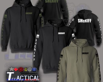 May include: A collection of hooded sweatshirts in black and olive green featuring the word SHERIFF printed on the chest and sleeves. Each hoodie includes a flag graphic on the sleeve. The image also displays the text TEEACTICAL with various emblems.