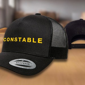 May include: Black and gray mesh trucker hat with a black brim and a yellow embroidered logo that says "CONSTABLE". The hat has a black snap closure and a label that says "The Classics Snapback Yupoong Since 1974".