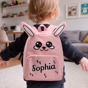 Könnte beinhalten: Ein rosa Kinderrucksack mit einem Hasengesicht-Design, einschließlich großer Ohren und einem niedlichen Ausdruck. Der Rucksack hat den Namen "Sophia" auf der Vorderseite aufgedruckt, zusammen mit Pfotenabdrücken.