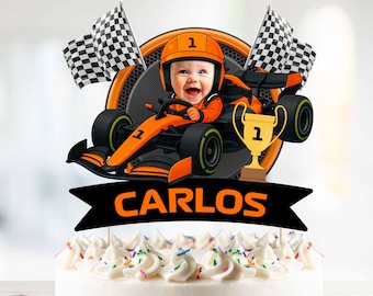 May include: A cake topper featuring a smiling baby in a race car, wearing an orange helmet. The car is orange and black, with a trophy and checkered flags. The name "CARLOS" is displayed on a black banner.