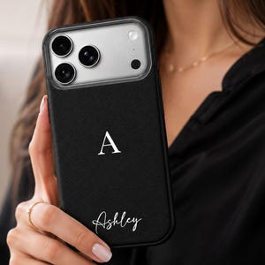 Könnte beinhalten: Eine schwarze Smartphone-Hülle mit strukturierter Oberfläche, die in der oberen Mitte mit einem großen weißen Serifen-Buchstaben A und im unteren Bereich mit dem Namen Ashley in weißer Schreibschrift verziert ist.