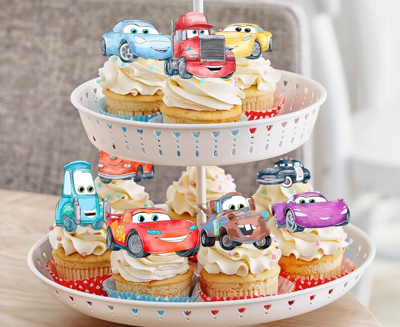 Puede incluir: Un soporte para pasteles blanco de dos niveles con cupcakes decorados con adornos de coches de dibujos animados. Los coches son rojos, azules, amarillos, morados y marrones. Los cupcakes est&aacute;n cubiertos con glaseado blanco y tienen chispas de colores.