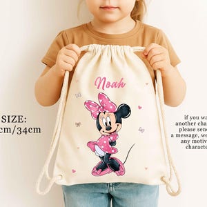 May include: A beige drawstring bag featuring a Minnie Mouse graphic in pink and black. The bag has the name "Noah" printed in pink, with dimensions of 16.5 inches by 13.4 inches. The bag also has a message about custom characters.