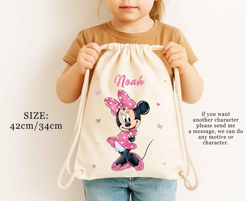 May include: A beige drawstring bag featuring a Minnie Mouse graphic in pink and black. The bag has the name "Noah" printed in pink, with dimensions of 16.5 inches by 13.4 inches. The bag also has a message about custom characters.