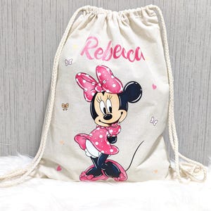 May include: A beige drawstring backpack featuring a Minnie Mouse graphic. Minnie is depicted in a pink polka-dot dress and bow. The name "Rebeca" is printed in pink script above the character. Small butterfly and bow illustrations are scattered around.
