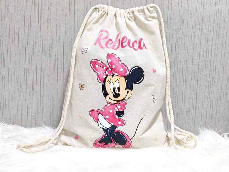 May include: A beige drawstring backpack featuring a Minnie Mouse graphic. Minnie is depicted in a pink polka-dot dress and bow. The name "Rebeca" is printed in pink script above the character. Small butterfly and bow illustrations are scattered around.