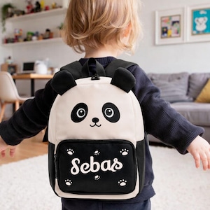 May include: A panda-themed backpack with black and white details. The backpack features a panda face design on the front, with the name "Sebas" printed in white script on the lower pocket. The backpack has black straps and a top handle.