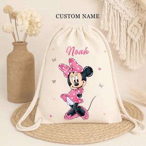 May include: A beige drawstring backpack featuring a Minnie Mouse graphic in pink and black. The name "Noah" is printed in pink, with the words "CUSTOM NAME" above. The bag has white drawstrings and small pink hearts and butterflies.