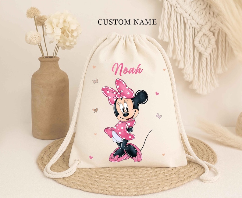 May include: A beige drawstring backpack featuring a Minnie Mouse graphic in pink and black. The name "Noah" is printed in pink, with the words "CUSTOM NAME" above. The bag has white drawstrings and small pink hearts and butterflies.
