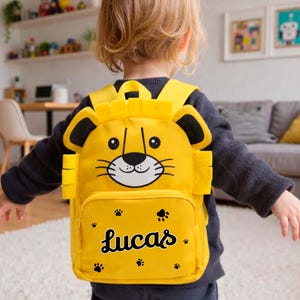 Op de afbeelding: Een gele rugzak met een leeuwengezicht ontwerp. De rugzak heeft een voorvak met de naam "Lucas" in een scriptlettertype en pootafdrukken. Het gezicht van de leeuw heeft zwarte en witte details. De rugzak heeft gele bandjes.