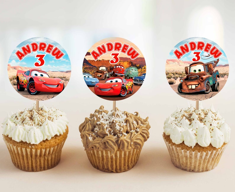 Pode incluir: Tr&ecirc;s cupcakes decorados com glac&ecirc; vermelho, branco e marrom. Cada cupcake tem um adesivo redondo e colorido com uma imagem de desenho animado de um carro de corrida vermelho, um carro de corrida azul e um guincho enferrujado. Os adesivos tamb&eacute;m t&ecirc;m o nome "Andrew" e o n&uacute;mero "3".
