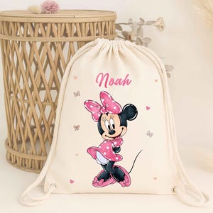 May include: A beige drawstring backpack featuring a Minnie Mouse graphic in a pink polka dot dress and bow. The name "Noah" is printed in pink above the character. Pink hearts and butterflies decorate the bag.