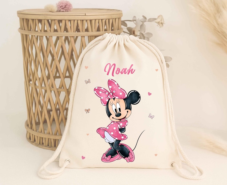 May include: A beige drawstring backpack featuring a Minnie Mouse graphic in a pink polka dot dress and bow. The name "Noah" is printed in pink above the character. Pink hearts and butterflies decorate the bag.