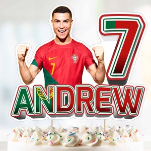 Puede incluir: Un adorno para pastel con un jugador de fútbol con una camiseta roja y verde con el número 7 en la parte delantera. El adorno también incluye el nombre "ANDREW" en letras verdes y rojas.