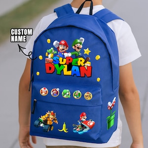 Op de afbeelding: Blauwe rugzak met Super Mario-personages, waaronder Mario, Luigi en Yoshi. De rugzak heeft een voorvak met karakterbadges en de naam "DYLAN" in kleurrijke letters. Een rode en witte paddenstoel staat aan de zijkant.