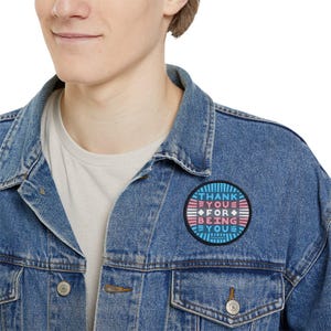 Op de afbeelding: Een persoon draagt een blauw denim jack met een ronde pin die "Dank je wel dat je jezelf bent" zegt in een blauw, roze en wit kleurenschema.
