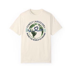 May include: Cream-colored t-shirt with a circular graphic featuring a green and blue globe, a blue ribbon, and the text "FAMILY CAREGIVERS MAKE THE WORLD GO ROUND" in blue and green. The shirt has short sleeves.