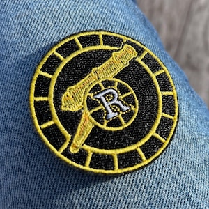 May include: Embroidered patch featuring a black and yellow circular design. The patch has a black background with a yellow border and a yellow crossed cannon. The center features a white letter "R".