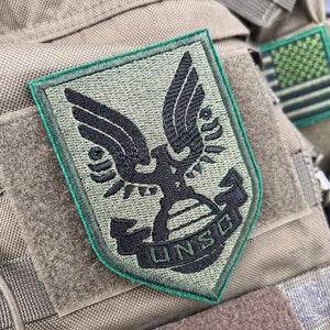 May include: A green and black embroidered patch with a stylized eagle design and the text "UNSC" in a black banner. The patch is attached to a brown fabric background.
