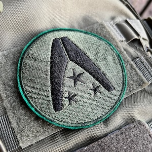 May include: A green and black embroidered patch with a stylized letter A and five stars. The patch is attached to a gray fabric background.