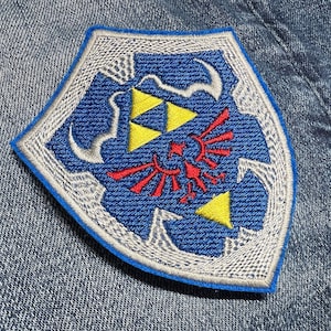 Puede incluir: Un parche bordado azul y blanco con el icónico símbolo de la Trifuerza de la serie de videojuegos The Legend of Zelda. La Trifuerza está rodeada por un diseño alado estilizado y un borde blanco.