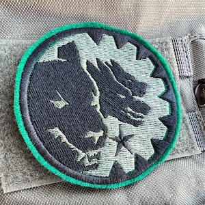 May include: A black and green embroidered patch with a stylized image of two wolves facing each other. The patch has a green border and is attached to a gray fabric background.