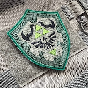 May include: A green and black embroidered patch featuring the Triforce symbol from the Legend of Zelda video game series. The patch is attached to a gray fabric background.